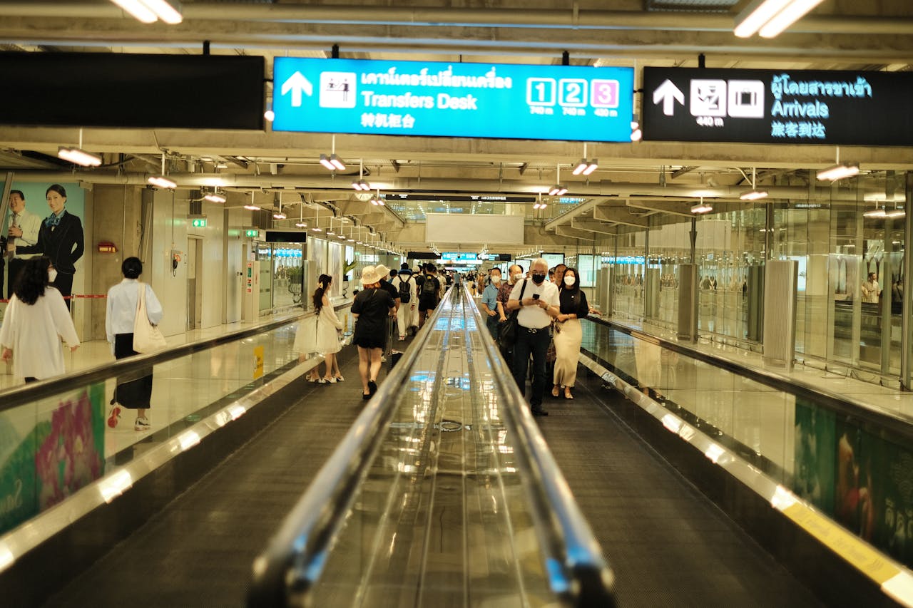 services-01 Passengers walk on a travelator in an airport arrival area, surrounded by signs and bright lighting.